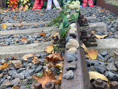 Foto von den Schienen beim Gedenkort Güterbahnhof Moabit auf denen einige Steine in Andenken an die Ermordeten liegen. Im Hintergrund sind die Gedenkkränze und einzelne weiße Rosen zu sehen.