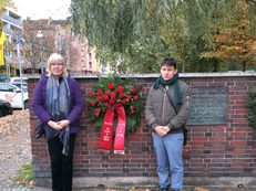 Die Bezirksverordneten Ingrid Bertermann und Leonard Diederich neben dem Gedenkkranz mit roten Blüten der Fraktion DIE LINKE. in der BVV Berlin-Mitte. Auf den Bändern steht der Name der Fraktion und: „Nie wieder – Ihr Schutz ist unsere Pflicht“. 