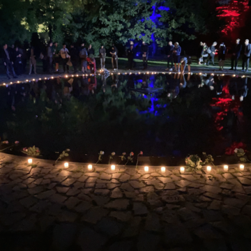 Ein Foto des zentralen Beckens des Mahnmals. Ringsherum wurden Kerzen angezündet und Blumen niedergelegt. Auf der anderen Seite stehen Menschen in Andacht und legen Blumen nieder.