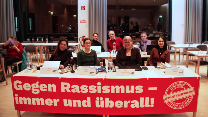 Ein Gruppenfoto der Linken in der BVV Mitte. An dem vorderen Tisch ist ein Banner angebracht auf dem steht „Gegen Rassismus – immer und überall!“. Zu sehen sind in der ersten Reihe von links nach rechts: Deniz Seyhun, Hannah Gerlof, Ingrid Bertermann und Jacky Sanehy. In der zweiten Reihe sitzen von links nach rechts: Samiullah Malekzadeh, Rüdiger Lötzer und Thilo Urchs. 