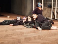 Bühnenfoto der  Gedenkaufführung im GRIPS-Theater. Zu sehen sind mehrere jungen Menschen mit Masken, die reglos auf dem Boden liegen, mit Ausnahme einer jungen Person mit blauer Maske, die aufrecht sitzt und ins Publikum blickt.  