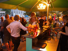 Foto von der Bar des Sommerempfang. Zu sehen sind mehrere kleine Grüppchen  und die Barkeeperin. Im Hintergrund steht eine DIE LINKE. Beachflag zwischen Palmen. 