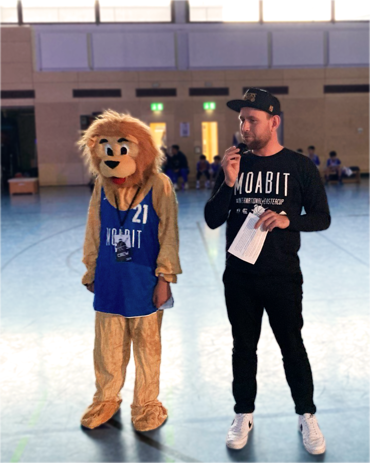 Foto von der Eröffnung des International Easter Cup Berlin Moabit 2024. Rechts zu sehen ist Jugendstadtrat Christoph Keller (Linke) beim Halten seiner Eröffnungsrede. Neben ihm steht eine Person in einem Löwenkostüm in einem Moabit 21 Trikot – das Maskottchen des Easter Cups.