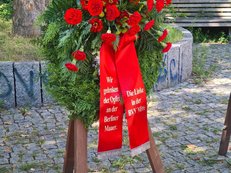 Foto des Kranzes der Linken in der Bezirksverordnetenversammlung Berlin Mitte in Gedenken an die Opfer der Berliner Mauer. Rote Rosen in einem grünen Kranz. Auf der linken Schleife steht „Wir gedenken den Opfern an der Berliner Mauer“. 