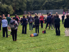 Foto von der Gedenkveranstaltung für den Porajmos am 2. August beim Mahnmal Tiergarten. Eine Gruppe Menschen steht und sitzt im Halbkreis um das zentrale Becken beim Mahnmal. Davor stehen Uwe Neumärker und Hamze Bytyçi während sie ihre Eröffnungsreden halten. 