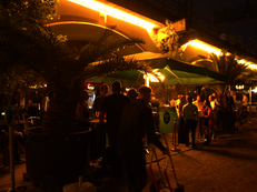 Foto vom Sommerempfang nach Sonnenuntergang. Mehrere Personengrupen stehen auf die Länge der Mokka Mitte Bar verteilt, die Leuchtröhren am S-Bahn Übergang tauchen die Szene in atmosphärisches Licht. 