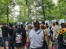 Foto vom Protest gegen die Entfernung des Mahnmals an den Porajmos in Berlin Tiergarten. Es ist eine große Gruppe Menschen zu sehen, die einem Redebeitrag vor den Gedenktafeln lauscht.