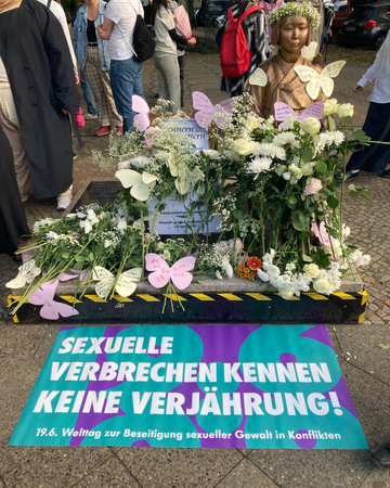 Ein Foto der Friedensstatue Ari in Berlin Moabit am 19. Juni. Sie ist bedeckt mit weiten Rosen und Zetteln mit Wünschen für die Zukunft. Auf einem Banner zu ihren Füßen steht: „Sexuelle Verbrechen kennen keine Verjährung! 19.6. Welttag zur Beseitigung sexueller Gewalt in Konflikten“