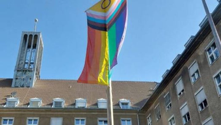 Foto der Progress-Pride Fahne während sie vor dem Rathaus Tiergarten gehisst wird vor einem blauem Himmel.