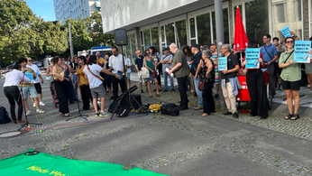 Foto der Kundgebung von BAVUL e.V. vor dem Rathaus Mitte. Im Hintergrund steht eine Gruppe von ca. 40 Menschen, vor ihnen ist ein grünes Banner ausgebreitet auf dem in Gelb „Hände Weg von Bavul e.V. für Kunst und Kultur – BAVUL“ steht. 