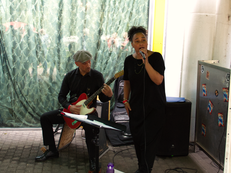 Foto vom Auftritt der Band Key of Life Pocket Soul Band. Eine Person singt in ein Mikrofon, links davon sitzt jemand und bedient ein elektronisches Schlagzeug Paddle während er auf einer elektrischen Gitarre spielt. 